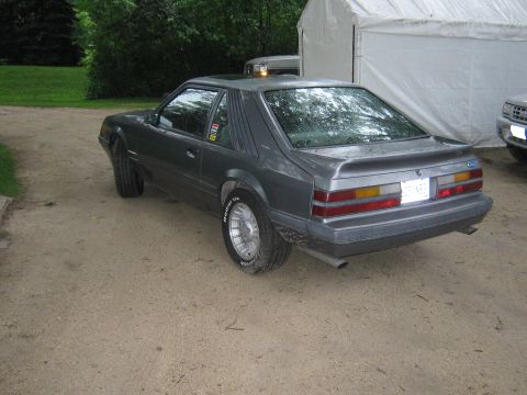 1985 Ford Mustang - A gray 1985 Ford Mustang hatchback is seen from a 3/4 front angle, parked on a dirt surface with a tent and another vehicle in the backgrou…