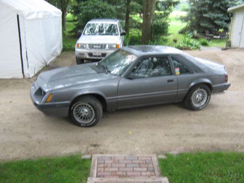 1985 Ford Mustang - A gray 1985 Ford Mustang is parked on a gravel driveway next to an SUV and a white tent.