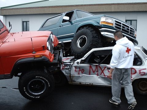 1996 Ford Bronco - A blue 1996 Ford Bronco is perched atop a mangled car, with a red Jeep positioned to its side.