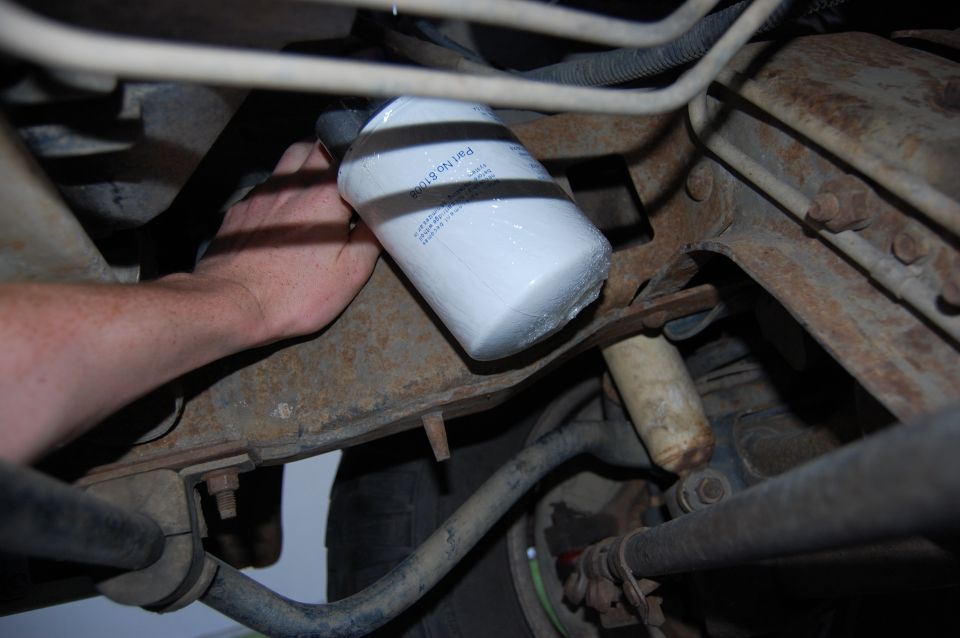 Hand installing white spin-on filter marked Part No 61008 under a truck near rusty frame and suspension parts