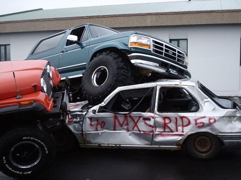 1996 Ford Bronco - A 1996 Ford Bronco is stacked on top of a wrecked sedan, with another vehicle partially visible.