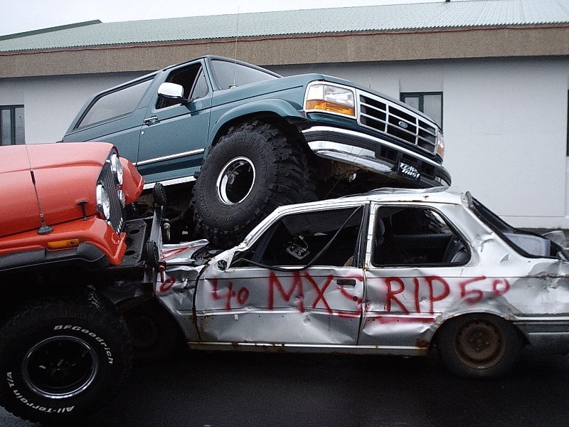 Green lifted Ford Bronco on oversized tires crushing a silver sedan tagged with red spray paint