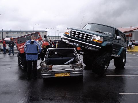 1996 Ford Bronco - A lifted Ford Bronco is parked on top of a damaged car, with a red Jeep in the background.