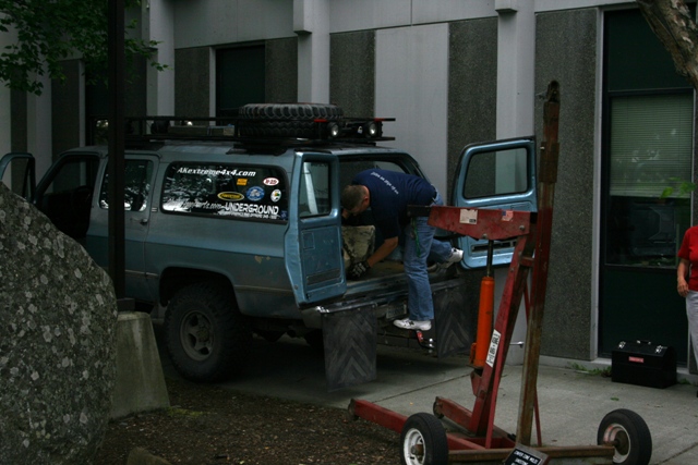 Blue SUV with roof rack and spare tire, person loading items into back. Red engine hoist nearby