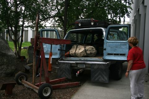1990 Chevrolet Suburban - A woman observes as a large rock is loaded into the back of a light blue SUV using a red engine hoist.