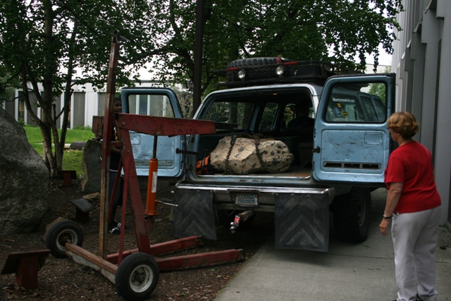 Light blue SUV with rear doors open, a large rock secured with chains inside, and a woman standing nearby. A red engine hoist is next to the vehicle