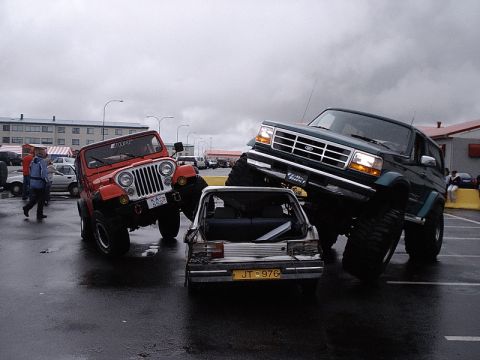1996 Ford Bronco - A modified Ford Bronco drives over a small car, with a red Jeep nearby.