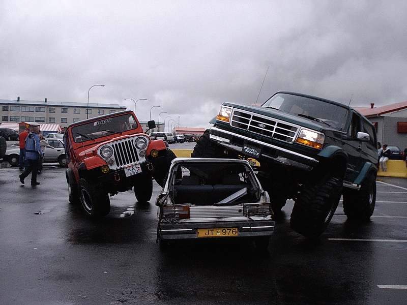 Lifted green Ford Bronco crushing silver car beside red Jeep in wet lot