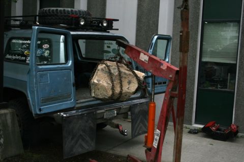 1990 Chevrolet Suburban - A rock is being loaded into the bed of a blue pickup truck using an engine hoist.