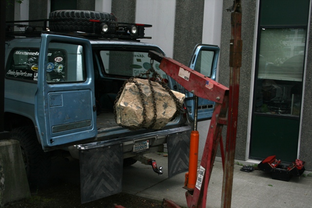 Blue pickup truck with doors open, a rock is being lifted into the bed by a red engine hoist