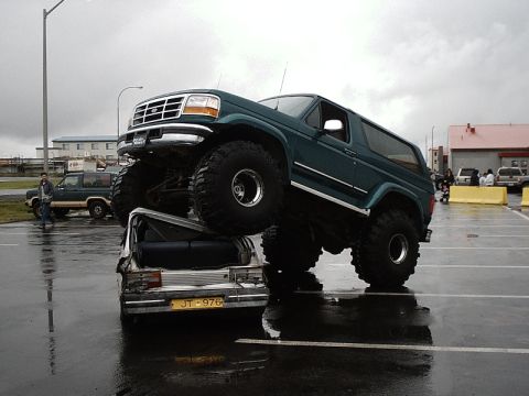 1996 Ford Bronco - A lifted 1996 Ford Bronco is parked on top of a wrecked car in a parking lot.