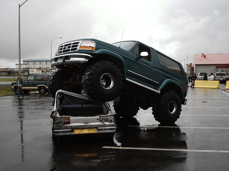 Lifted teal Ford Bronco on huge tires parked atop a crushed white sedan in a wet lot