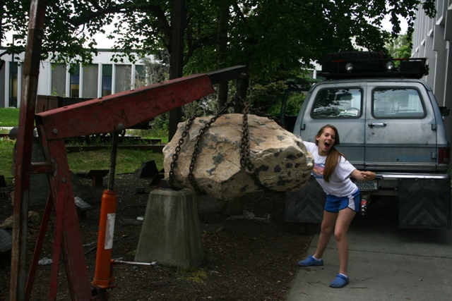 Girl posing with a large rock being lifted by a red crane next to a blue van