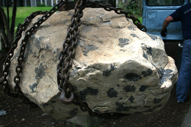 Large rock suspended by chains, with a blue vehicle and person in the background