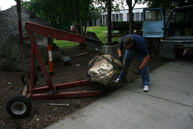 Engine hoist lifting a large rock with a man securing straps. Blue truck in background