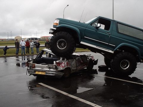 1996 Ford Bronco - A lifted 1996 Ford Bronco is parked on top of a severely damaged car, with onlookers observing the scene.