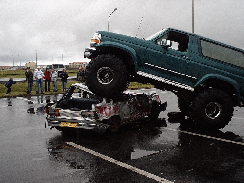 Lifted teal Ford Bronco on big tires crushing a silver car in a wet lot as spectators watch