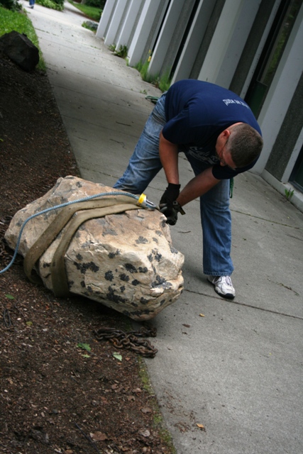 1990 Chevrolet Suburban - A person is using straps and chains to move a large boulder next to a building.