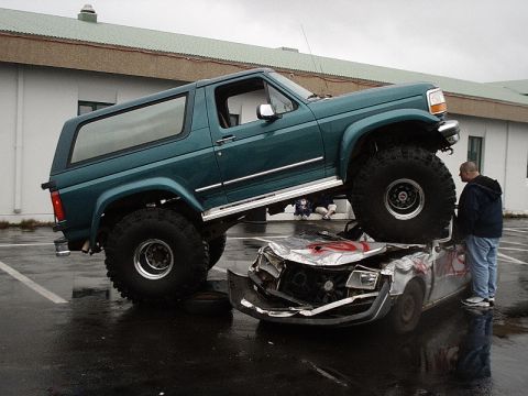 1996 Ford Bronco - A large green SUV is parked on top of a severely damaged car in a parking lot.