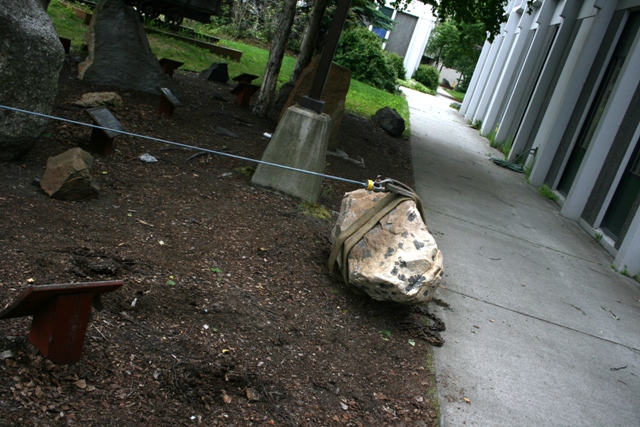 Rock secured with straps and cable on dirt next to a concrete path and building