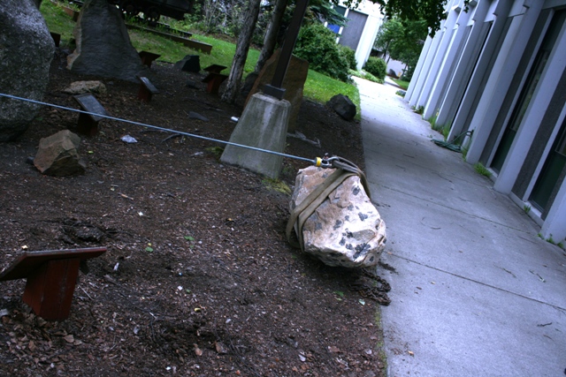 Rock with straps and cable attached, on dirt next to a concrete path and building