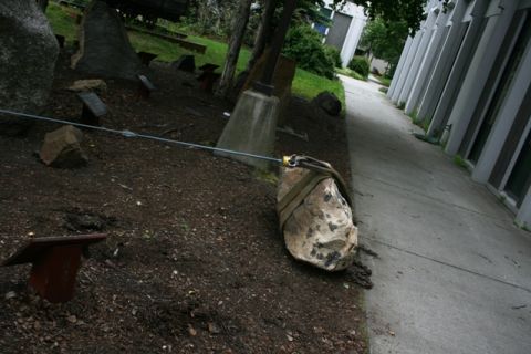 1990 Chevrolet Suburban - A large stone is held in place by straps and cables within a rock garden setting, adjacent to a modern building.