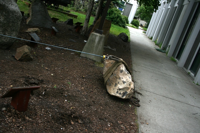 Rock secured with a strap and cable, resting on dirt next to a concrete path and building