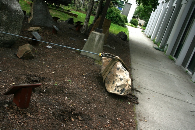 Rock with straps and cable on dirt path next to concrete walkway and building