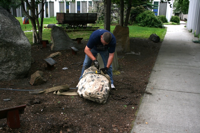 Man straps large rock for moving, with chains and a mining cart in background