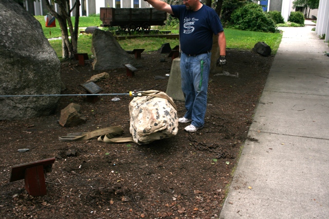 Man in blue shirt and jeans stands by a large, speckled rock with straps and chains on the ground
