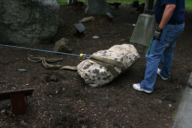 Man in jeans and gloves uses a strap and winch to move a large, rough rock on the ground