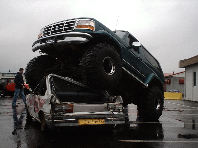 Lifted green Ford Bronco on big tires parked atop a crushed white sedan in a wet lot