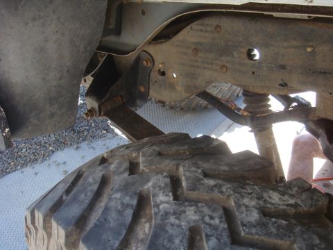 Close-up of a large, knobby tire and rusty suspension components of a vehicle. A person's leg is visible in th