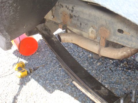 Close-up of vehicle frame, leaf spring, and exhaust pipe. Orange bucket and yellow drill visible