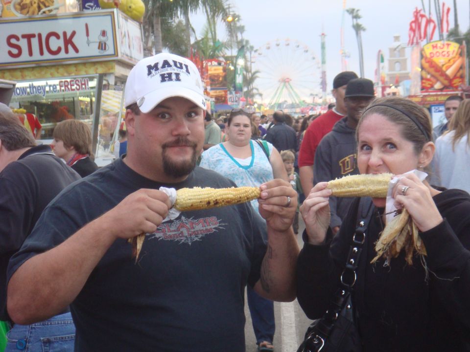 Man and woman eating corn on the cob at a fair with rides in the background
