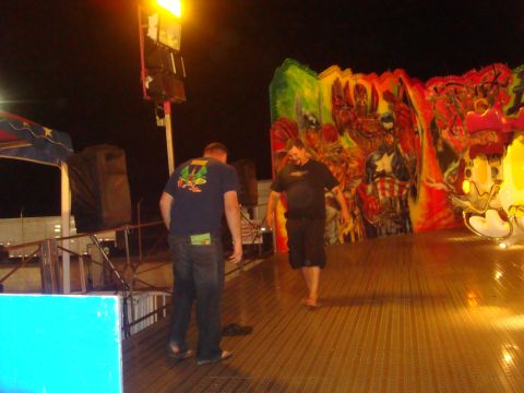 Two men on a carnival ride platform with a mural of superheroes painted on the wall behind them. "SUPERMOTORS.