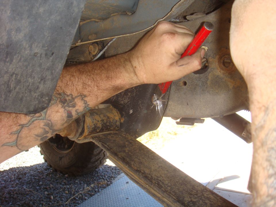 Close-up of a person's tattooed arm working on vehicle underside with a red tool.