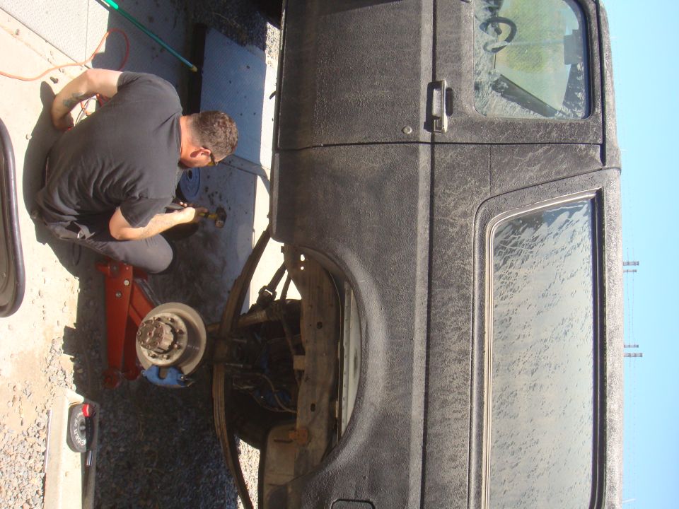 Man kneeling, working on the front axle of a dark-colored vehicle supported by a red jack