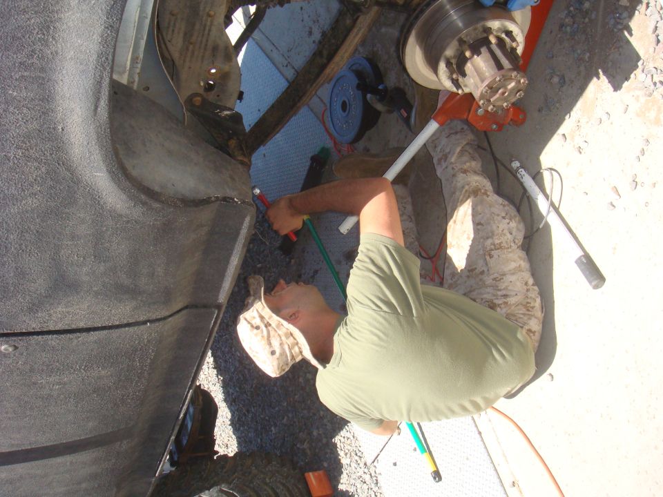 Person in camouflage pants and green shirt working on a vehicle's axle with tools