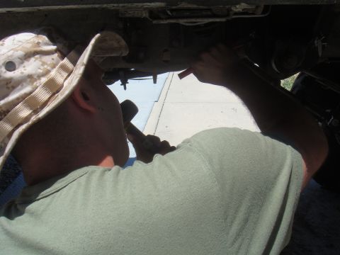 Person in camouflage hat and green shirt working on vehicle undercarriage with tools