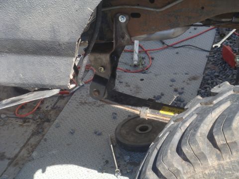 Close-up of a vehicle's leaf spring, shock mount, and part of a large tire. Yellow sticker on spring reads 'PO