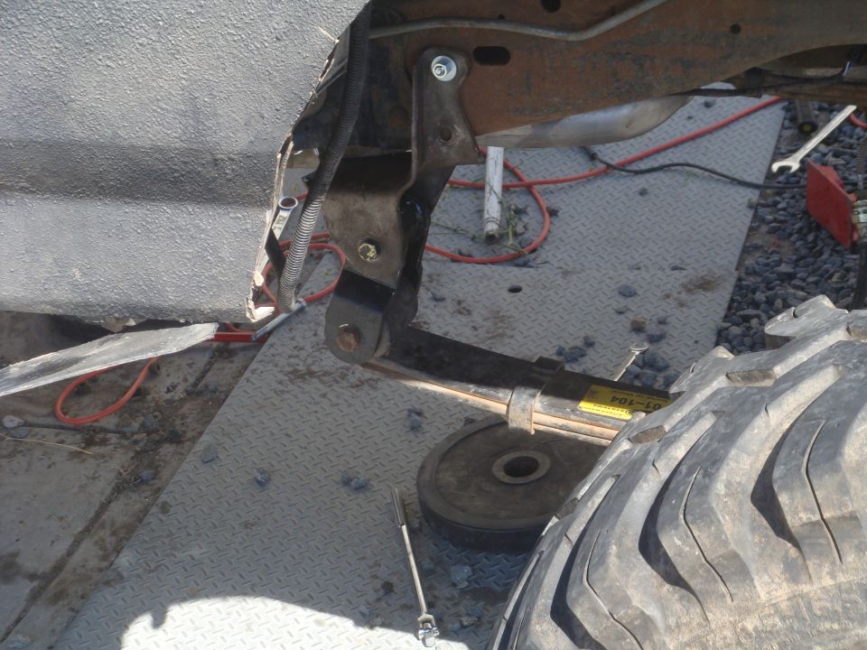 Close-up of a vehicle's leaf spring, shock mount, and part of a large tire. Yellow sticker on spring reads 'POS-ED'