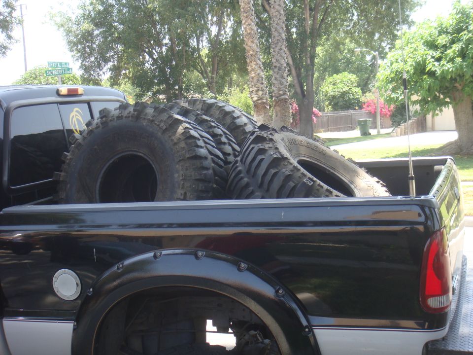 Black pickup truck bed loaded with four large off-road tires. Riveted fender flares visible