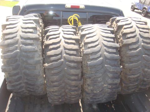 Four large off-road tires loaded in the bed of a pickup truck. A blue Jeep is visible in the background