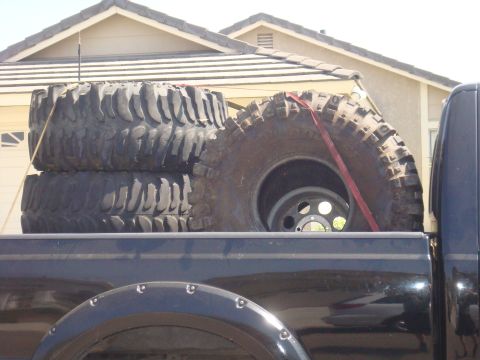 Black truck bed loaded with three large off-road tires, one strapped down