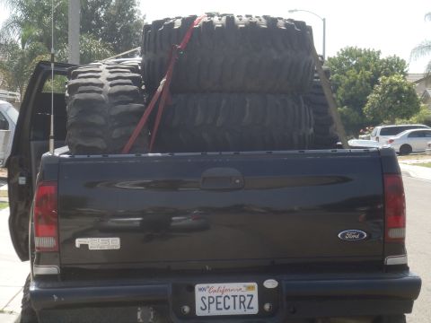 Rear view of black Ford F-250 pickup truck bed loaded with four large off-road tires. License plate reads SPEC