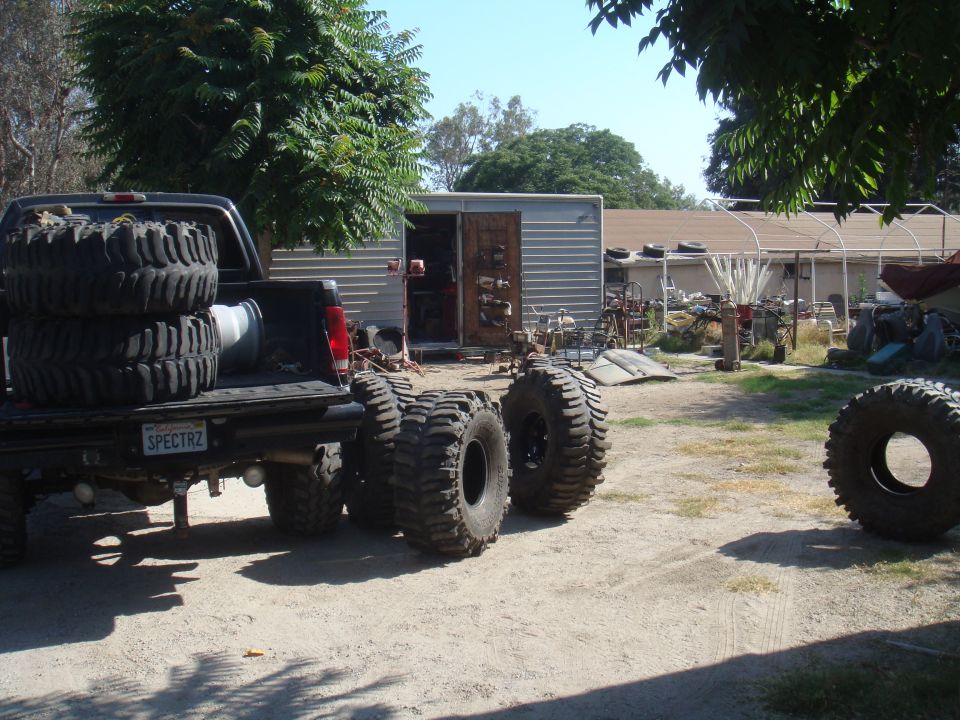 Pickup truck bed loaded with large tires. More tires are on the ground next to it. Yard has a shed and other items