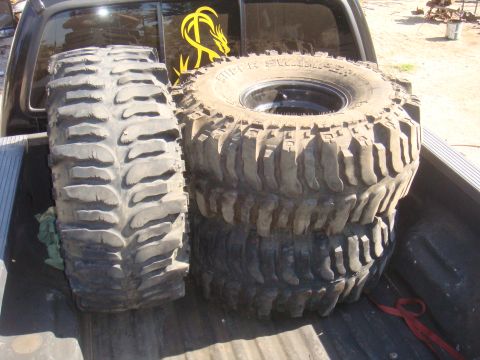 Truck bed holding four large off-road tires, one standing vertically and three stacked horizontally. A yellow