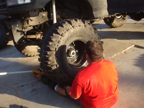 Man in red shirt working on a massive off-road tire mounted on a vehicle. The vehicle's suspension and front b