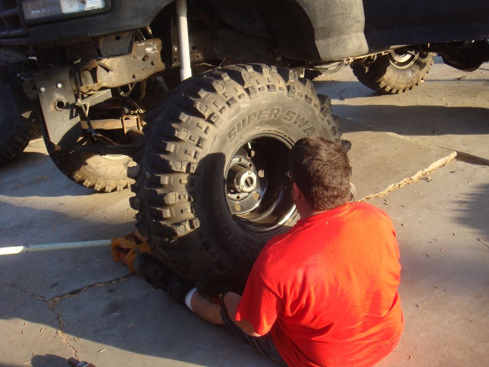 Man in red shirt working on a massive off-road tire mounted on a vehicle. The vehicle's suspension and front bumper are visible